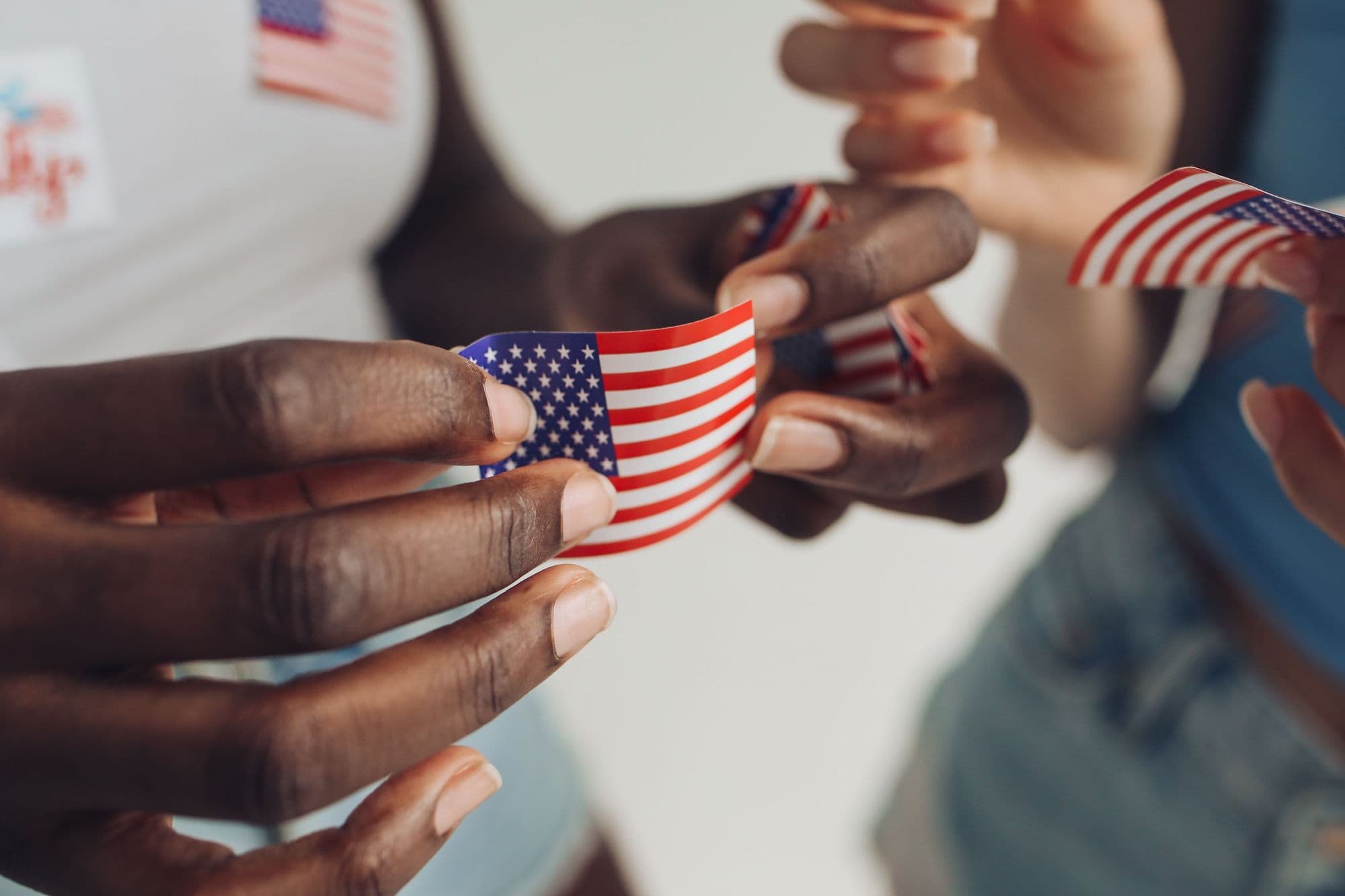 Several hands of different skin tones hold small American flags together.