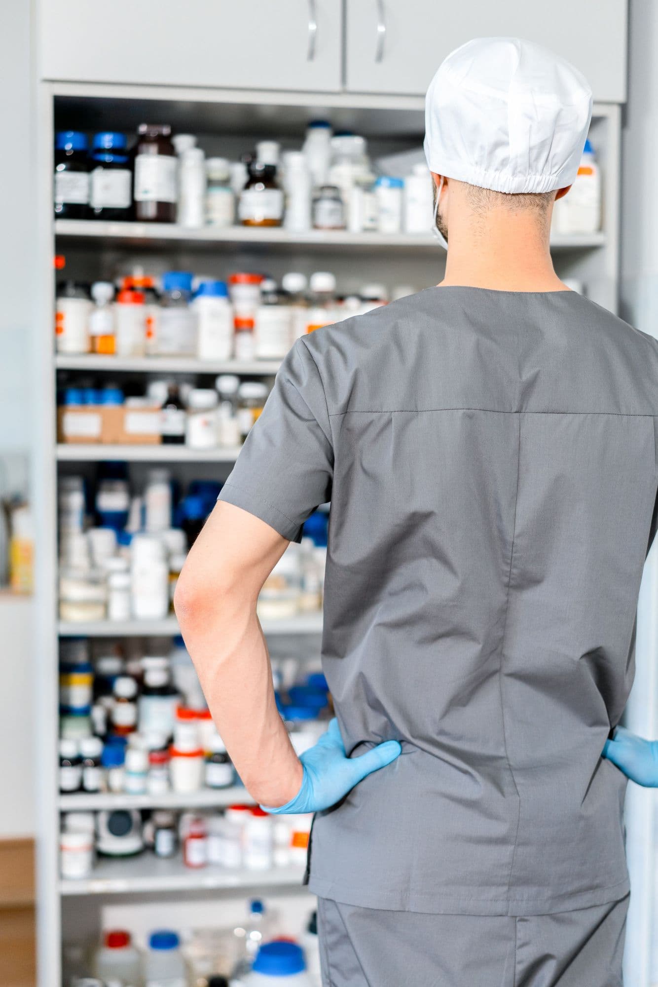 A pharmacy team member in scrubs and gloves looks over well-organized medication shelves.