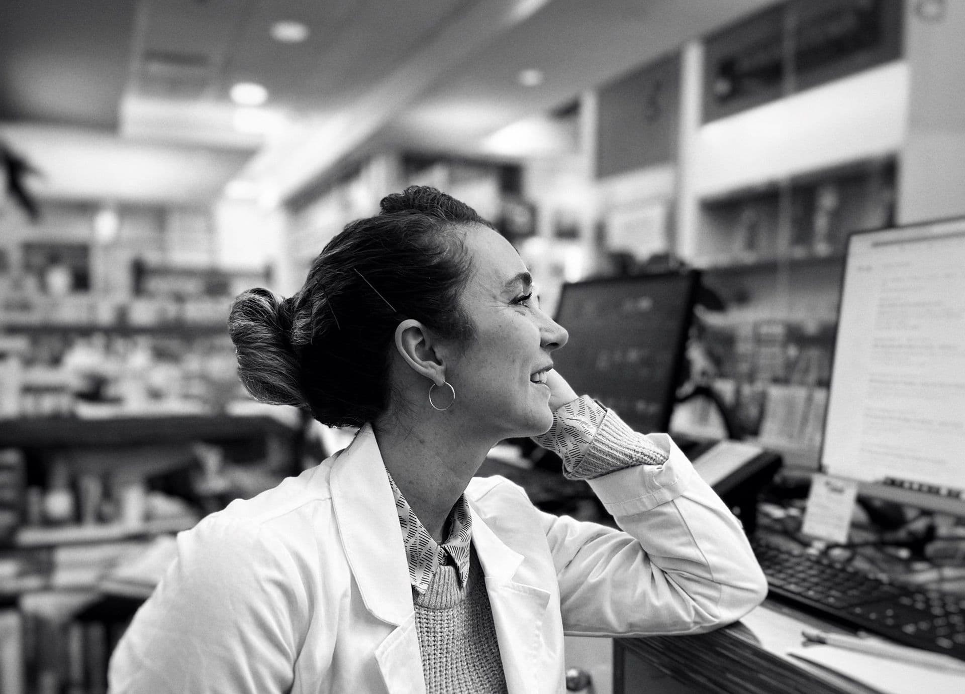 Black-and-white photo of a smiling pharmacist leaning on the dispensary counter.