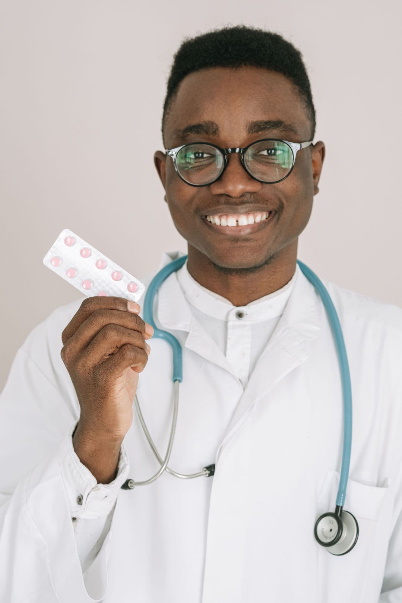 A smiling pharmacist with glasses and a stethoscope holds up a blister pack.