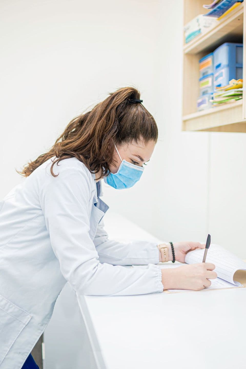 A pharmacy technician in a white coat writes notes at a bright workstation.